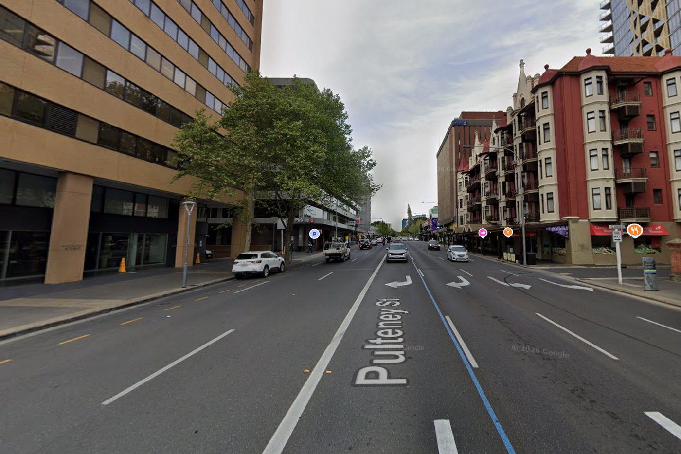 The same view in 2026: five lanes of asphalt, turning arrows, parked cars and a four-wheel drive, with the Ruthven Mansions still on the right.