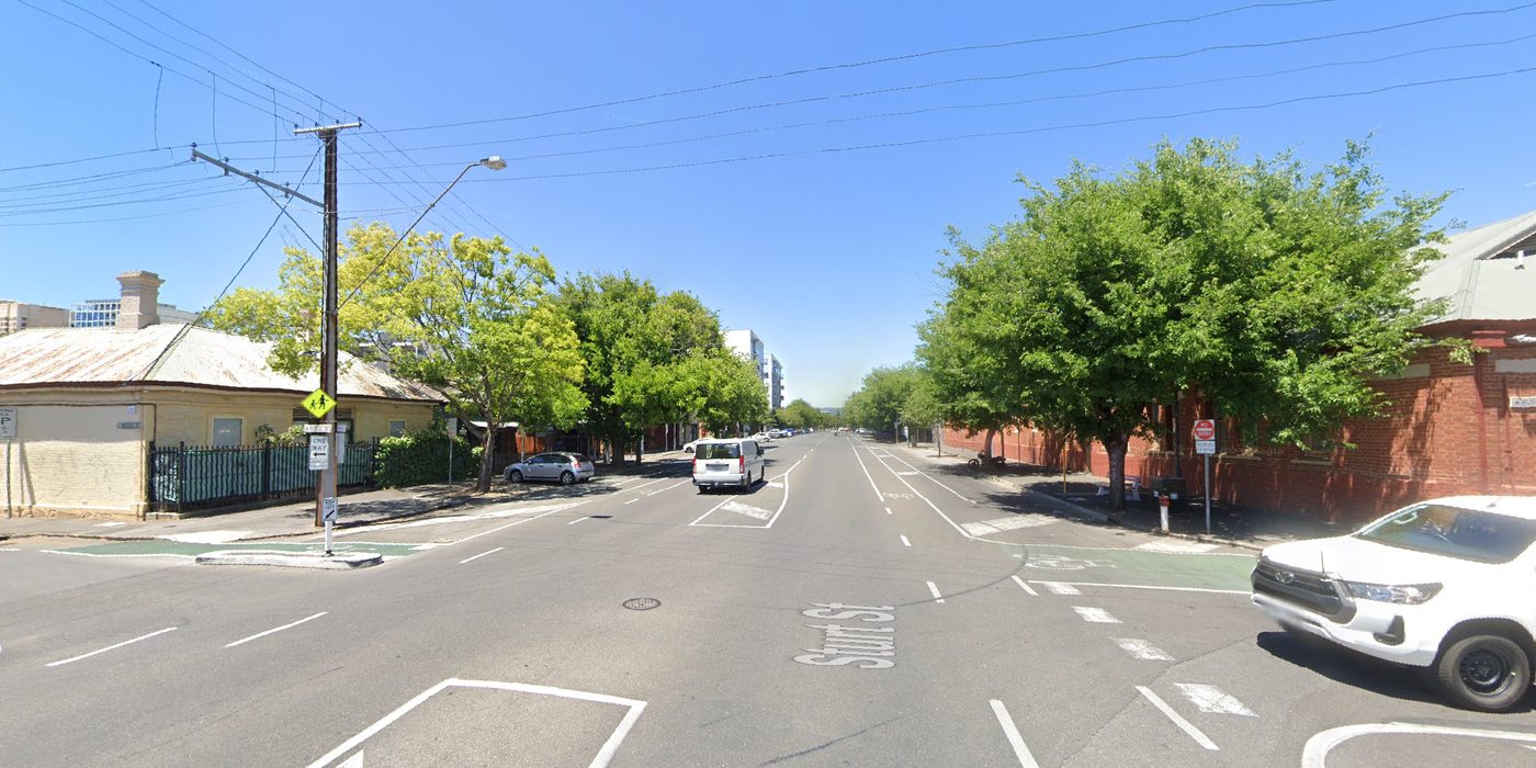 Sturt Street at Russell Street today, looking east: four lanes of asphalt, painted islands and turning arrows, a school-zone sign, street trees, and old brick and stone buildings on both corners.